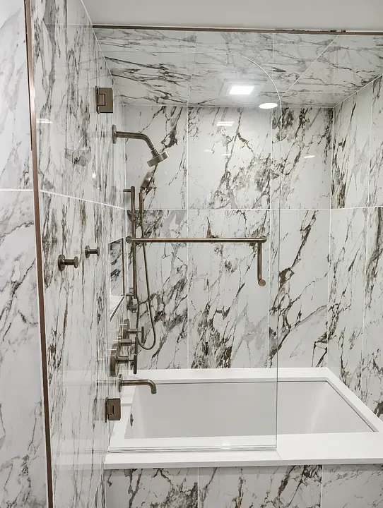 Marble-tiled shower with glass door, metal fixtures, and recessed lighting above a white bathtub.