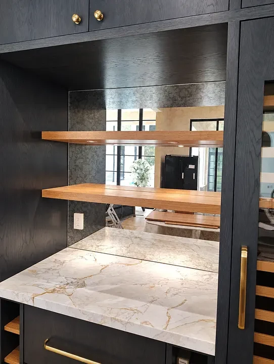 Modern kitchen bar with marble countertop, mirrored backsplash, wooden shelves, and dark cabinetry with brass accents.
