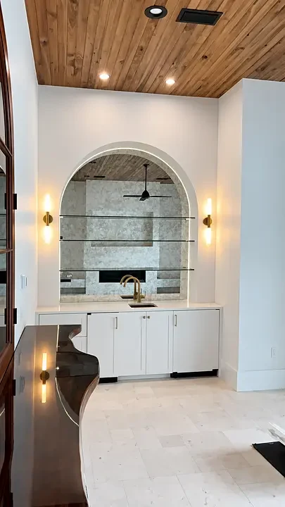 Modern kitchen with archway, glass shelves, wood ceiling, white cabinets, and brass faucet. Warm lighting on walls.