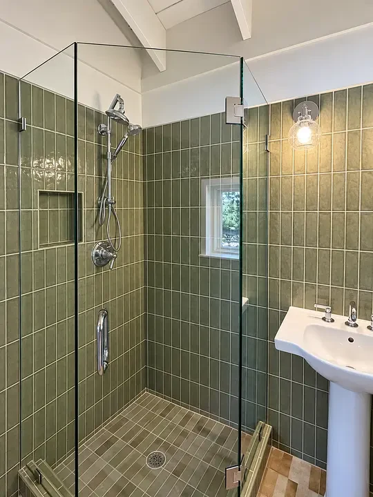 A glass shower with green tiles beside a white sink and a round light fixture on the wall.