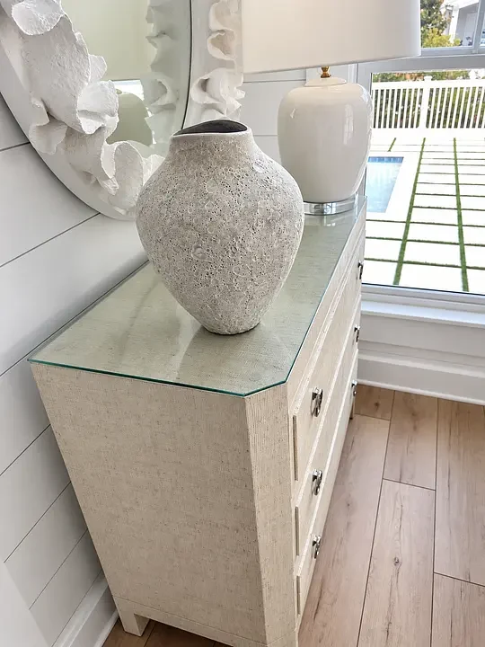 Textured vase on a glass-topped dresser, with a white lamp and mirror, beside a window overlooking a pool.