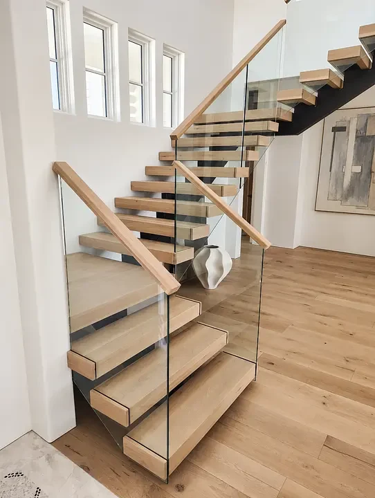 Modern wooden staircase with glass railing, light wood steps, and minimalist design in a bright room with windows.