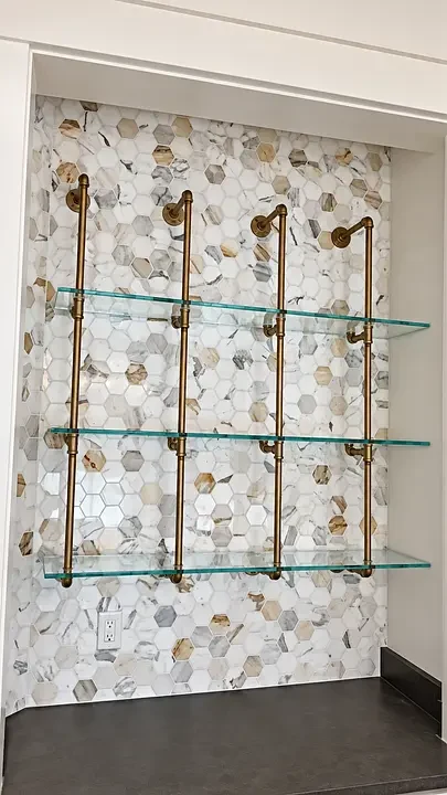 Glass shelves with brass supports on a hexagonal marble tile wall, above a dark countertop.