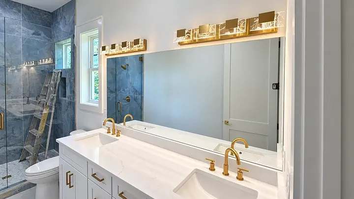Modern bathroom with dual sinks, gold fixtures, large mirror, blue-tiled shower, and a ladder beside the toilet.